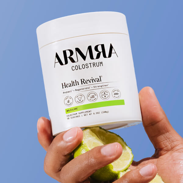 Hand holding a container of ARMRA Colostrum with limes against a blue background